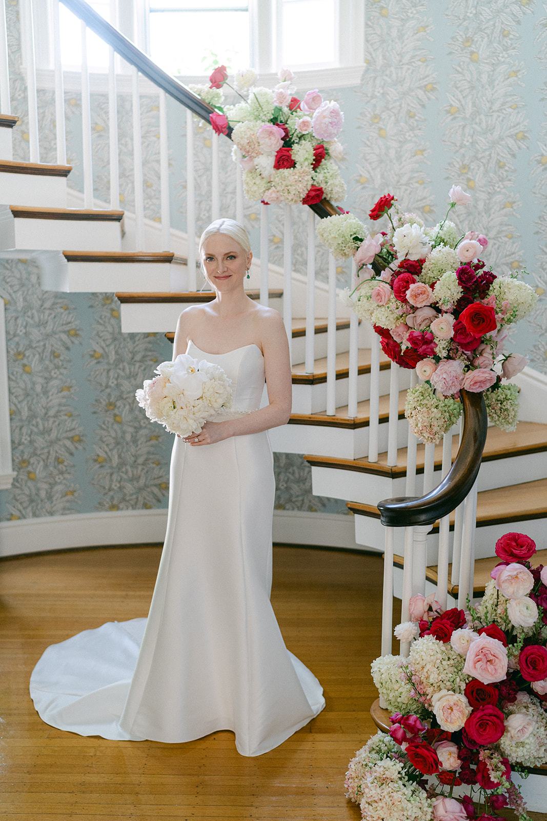 Bride Lauren standing by the staircase at her childhood home in Sewickley, Pennsylvania before her late-summer wedding, surrounded by lust pink floral arrangements and garden-inspired blooms.