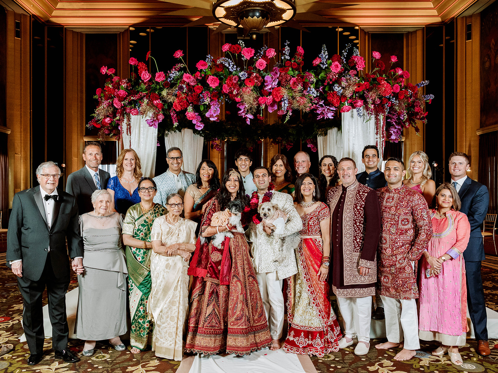 Family portrait beneath a floral mandap at an Indian wedding reception in the Urban Room at The Omni William Penn Hotel in Pittsburgh, with the couple's dogs Stevie and Otis included in the celebration.